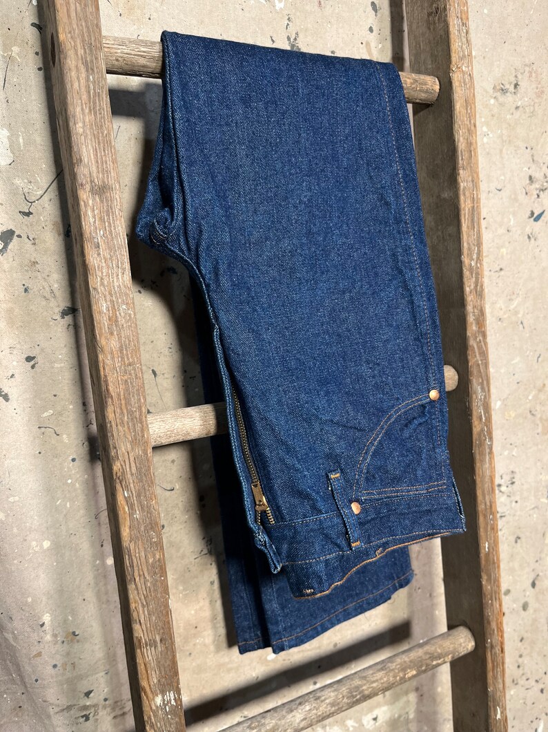 Puede incluir: Un par de pantalones vaqueros de mezclilla azul oscuro con cierre de cremallera y trabilla de cintur&oacute;n de cuero marr&oacute;n. Los vaqueros est&aacute;n colgados de una escalera de madera.