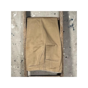 May include: Tan-colored pants folded over a wooden ladder. The pants have a classic design with back pockets and a button closure. The background is a concrete surface with paint splatters.