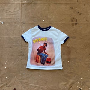 Pode incluir: Camiseta branca com uma gola azul, com um gráfico de um homem sentado em uma caixa segurando um pavio de dinamite aceso. O gráfico diz "Dyn-O-mite".
