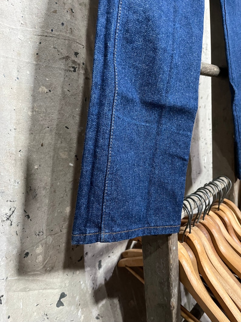May include: A pair of dark blue denim jeans with a straight leg, hanging on a wooden rack.