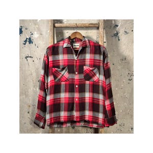 May include: A long-sleeved plaid shirt in shades of red, black, and gray, with two front pockets. The shirt is hanging on a wooden hanger against a neutral background. The shirt has a classic, button-down design.