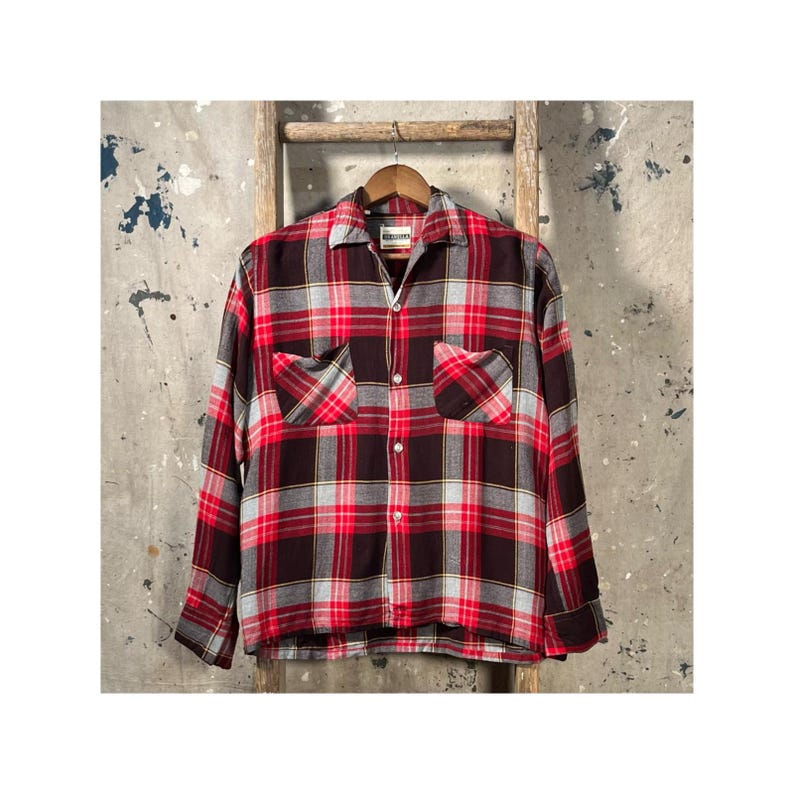 May include: A long-sleeved plaid shirt in shades of red, black, and gray, with two front pockets. The shirt is hanging on a wooden hanger against a neutral background. The shirt has a classic, button-down design.