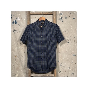 Könnte beinhalten: Kurzärmeliges Button-Down-Hemd in einem marineblauen und goldfarbenen Karomuster. Das Hemd hängt an einem Holzbügel. Das Hemd hat einen Button-Down-Kragen und einen gebogenen Saum.