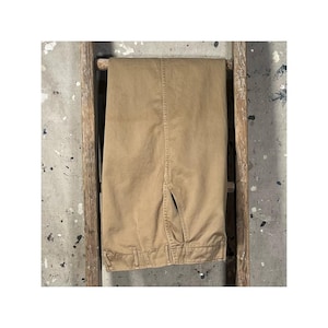May include: Khaki-colored pants hanging on a wooden ladder. The pants are neatly folded, showcasing the front pockets and belt loops. The background is a concrete wall with paint splatters.