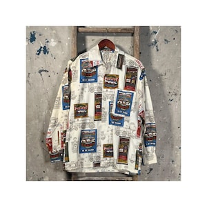 Op de afbeelding: Wit overhemd met knopen met een herhaalde print van vintage treinadvertenties in rood, blauw en bruin. Het shirt heeft lange mouwen en een kraag. Het ontwerp bevat de tekst "Pacific" en "Good Acres".