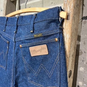May include: A pair of dark blue denim jeans with a leather Wrangler patch on the back pocket. The jeans are hanging on a wooden hanger.