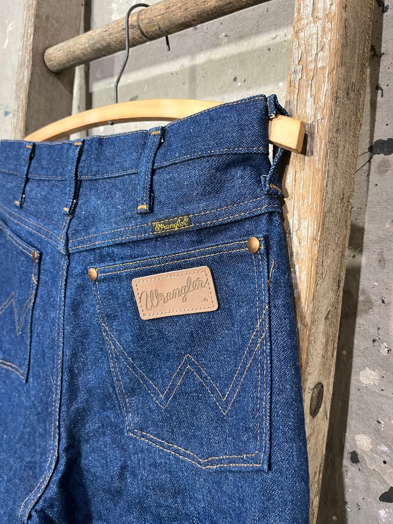 May include: A pair of dark blue denim jeans with a leather Wrangler patch on the back pocket. The jeans are hanging on a wooden hanger.