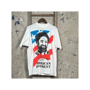 Puede incluir: Camiseta blanca con un diseño de bandera americana roja, blanca y azul. La bandera está detrás de una imagen en blanco y negro de un hombre con barba y una firma que dice "Bell Biv Devoe". El texto "AMERICAN DISSIDENT" está impreso debajo de la imagen.