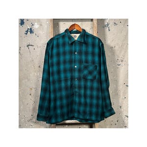 Pode incluir: Camisa de manga comprida xadrez verde-azulado e preto, com fecho de botões e bolso no peito. A camisa está pendurada num cabide de madeira. O fundo é uma parede neutra e texturizada.