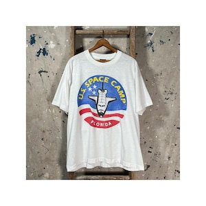 May include: White t-shirt featuring a graphic of a space shuttle inside a blue circle with the text "U.S. SPACE CAMP FLORIDA". The design includes red and white stripes. The shirt is hanging on a wooden hanger.