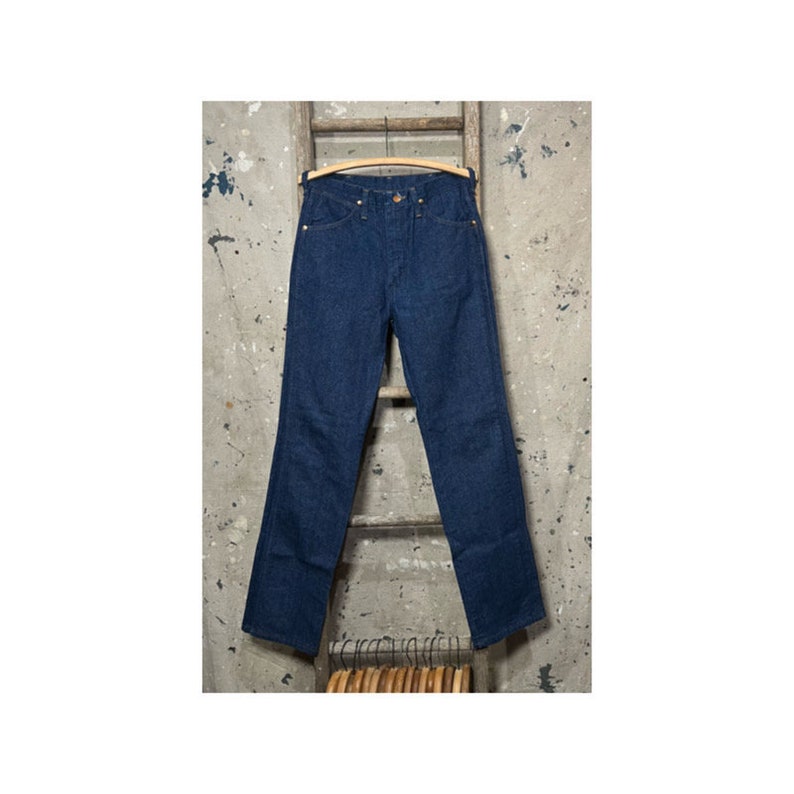 May include: A pair of dark blue denim jeans hanging on a wooden ladder. The jeans have a classic straight leg style and a button fly.