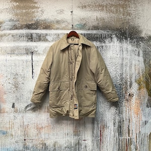 Puede incluir: Una chaqueta beige de manga larga con cierre de botones y cintura con cordón. La chaqueta tiene dos bolsillos grandes en la parte delantera y un pequeño bolsillo de parche en el pecho izquierdo.