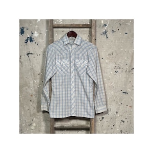 Puede incluir: Una camisa de manga larga de cuadros blancos y azules con cuello puntiagudo y dos bolsillos en el pecho. La camisa tiene un cierre de botones en la parte delantera y está colgada de una escalera de madera.