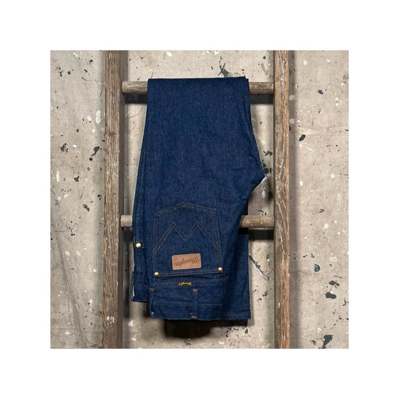Puede incluir: Un par de pantalones vaqueros de mezclilla azul oscuro colgados de una escalera de madera. Los pantalones tienen un parche de cuero marr&oacute;n con la palabra "Wrangler" en &eacute;l.