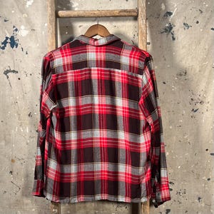 May include: A long-sleeved, button-down shirt with a classic plaid pattern in red, black, gray, and yellow. The shirt is hanging on a wooden hanger against a neutral background, showcasing the back of the garment.