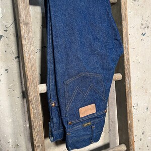Puede incluir: Un par de pantalones vaqueros de mezclilla azul oscuro colgados de una escalera de madera. Los pantalones tienen un parche de cuero en el bolsillo trasero con el texto "Wrangler" en &eacute;l.