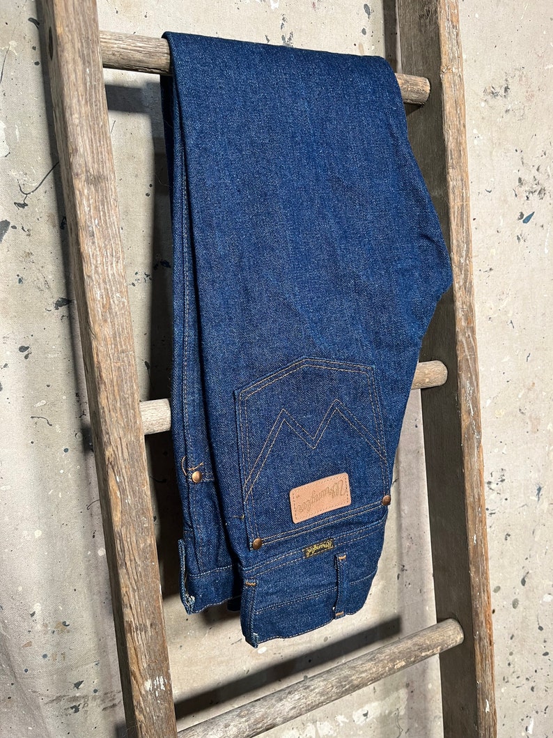 Puede incluir: Un par de pantalones vaqueros de mezclilla azul oscuro colgados de una escalera de madera. Los pantalones tienen un parche de cuero en el bolsillo trasero con el texto "Wrangler" en &eacute;l.