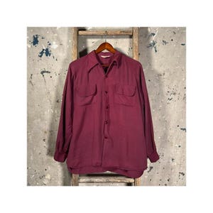 May include: A long-sleeve button-down shirt in a deep burgundy color. The shirt features a classic collar, two chest pockets with flaps, and a front button closure. The sleeves are long with buttoned cuffs, and the fabric has a smooth texture.