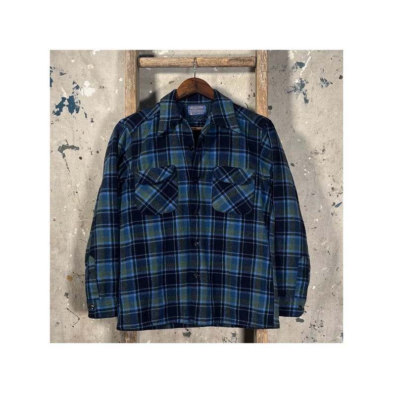 May include: A blue, green, and black plaid flannel shirt with two chest pockets and a collared neckline. The shirt is hanging on a wooden hanger against a neutral background.