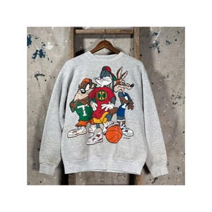 Puede incluir: Una sudadera gris claro de cuello redondo con un estampado gráfico de tres personajes de dibujos animados. El personaje central es un conejo con camiseta y gorra rojas, flanqueado por una criatura marrón con camiseta verde y un coyote azul.