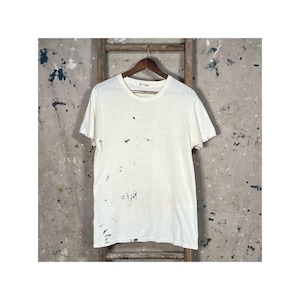 May include: Cream-coloured, short-sleeved t-shirt with black paint marks. The shirt is suspended on a wooden hanger against a distressed wooden ladder and a mottled grey backdrop.