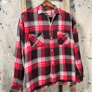 May include: A red, black, and gray plaid button-down shirt with two chest pockets. The long-sleeved shirt is hanging on a wooden hanger against a rustic background. The shirt has a classic collar and buttoned cuffs.