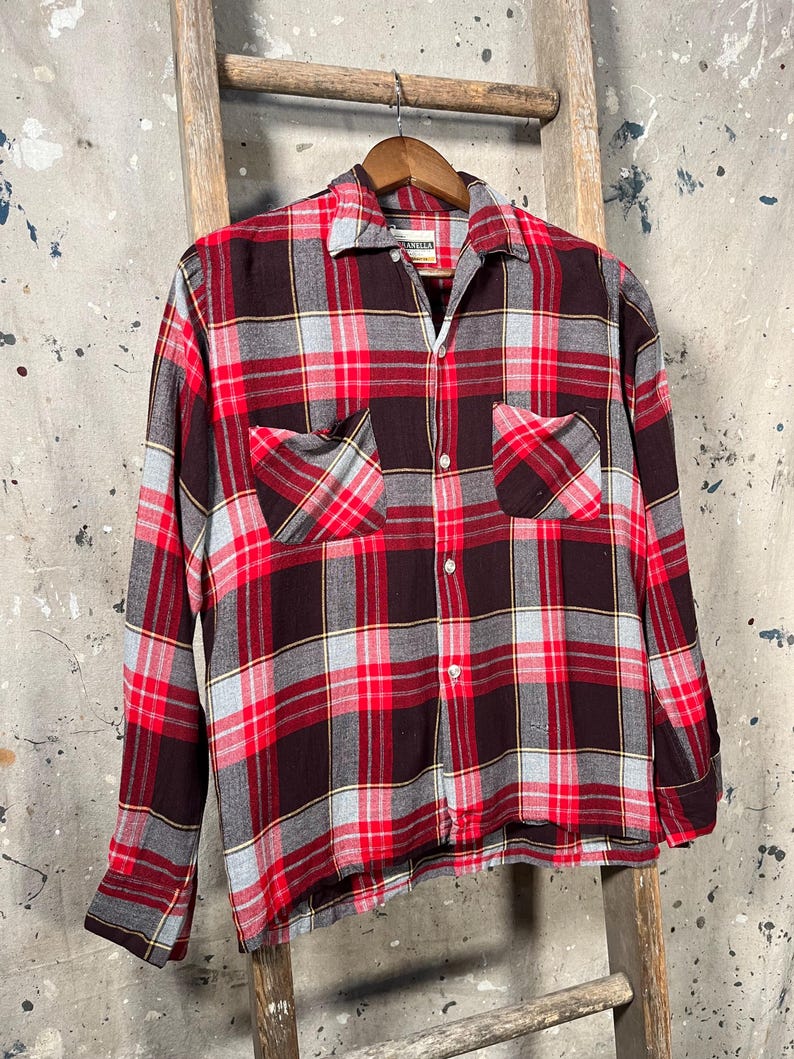May include: A red, black, and gray plaid button-down shirt with two chest pockets. The long-sleeved shirt is hanging on a wooden hanger against a rustic background. The shirt has a classic collar and buttoned cuffs.