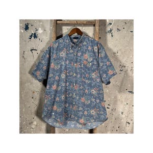 May include: A short-sleeved button-down shirt in a blue floral pattern. The shirt features a classic collar and a single chest pocket. The floral design includes pink, yellow, and green flowers on a blue background. The shirt is hanging on a wooden hanger.