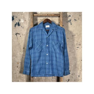 Puede incluir: Una camisa azul de manga larga a cuadros con dos bolsillos en el pecho y cuello de campamento. La camisa es abotonada y cuelga de una percha de madera. El fondo es una pared neutra y texturizada con una escalera de madera.