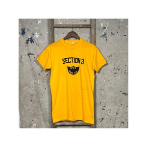 Può includere: Una maglietta gialla con una grafica in bianco e nero che dice "SECTION 3" e "NYS  PHS AA".
