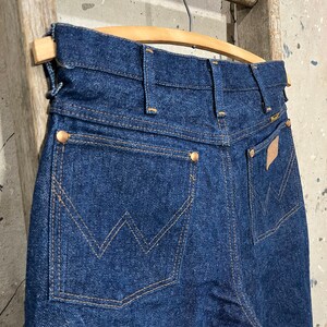 May include: A pair of dark blue denim jeans with a back pocket featuring a stitched W logo. The jeans are hanging on a wooden hanger.