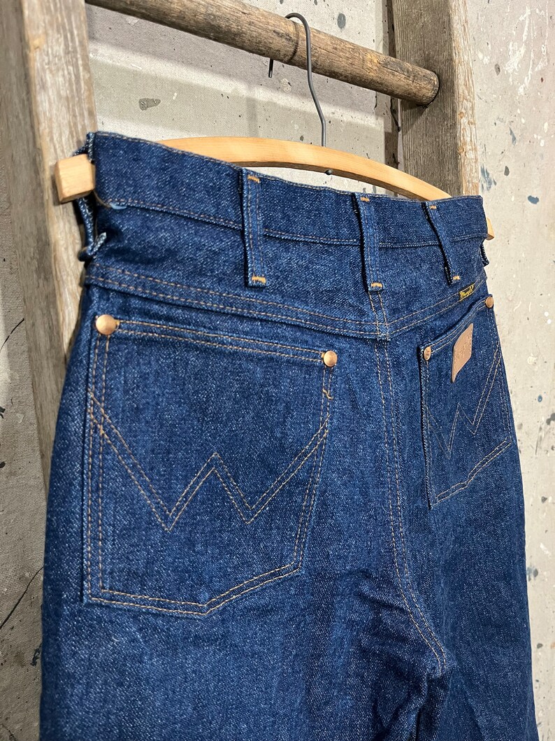 May include: A pair of dark blue denim jeans with a back pocket featuring a stitched W logo. The jeans are hanging on a wooden hanger.