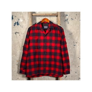 Puede incluir: Una camisa de franela a cuadros rojos y negros con mangas largas. La camisa tiene dos bolsillos en el pecho y una botonadura frontal. Cuelga de una percha de madera sobre un fondo neutro.