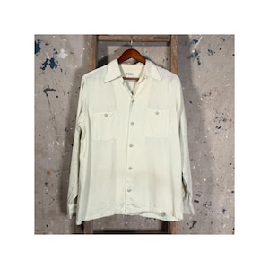 Puede incluir: Una camisa verde claro de manga larga con dos bolsillos en el pecho y cuello. La camisa está abotonada por delante y cuelga de una percha de madera. El fondo es una pared neutra y texturizada.