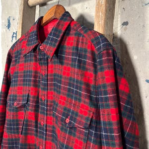 Puede incluir: Una camisa de franela a cuadros rojos, azul marino y verdes con dos bolsillos en el pecho y botones rojos. La camisa est&aacute; colgada de una percha de madera. La camisa tiene un estilo cl&aacute;sico y vintage.