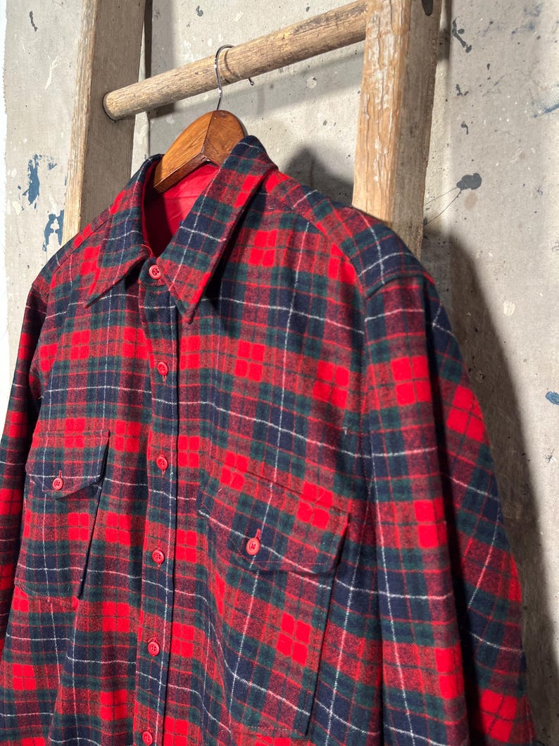 Puede incluir: Una camisa de franela a cuadros rojos, azul marino y verdes con dos bolsillos en el pecho y botones rojos. La camisa est&aacute; colgada de una percha de madera. La camisa tiene un estilo cl&aacute;sico y vintage.