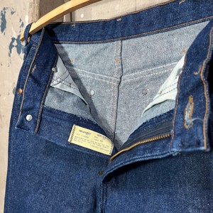 May include: A pair of dark blue denim jeans with a zipper fly and a Wrangler label. The label reads "Wrangler,  Mens Slim Fit,  100% Cotton,  Made in USA,  Size 32,  Waist 32,  Inseam 32,  Care Instructions: Machine Wash Cold, Tumble Dry Low,  Do Not Bleach,  Do Not Iron,  Do Not Dry Clean."