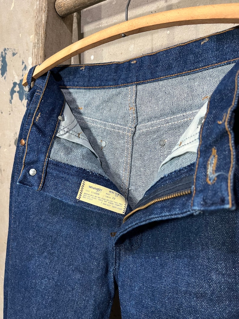 May include: A pair of dark blue denim jeans with a zipper fly and a Wrangler label. The label reads "Wrangler,  Mens Slim Fit,  100% Cotton,  Made in USA,  Size 32,  Waist 32,  Inseam 32,  Care Instructions: Machine Wash Cold, Tumble Dry Low,  Do Not Bleach,  Do Not Iron,  Do Not Dry Clean."