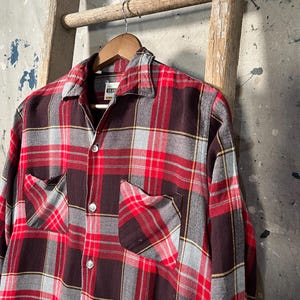 May include: A red, black, and gray plaid flannel shirt with two chest pockets and button closures. The shirt is hanging on a wooden hanger. The shirt has a classic, vintage style.