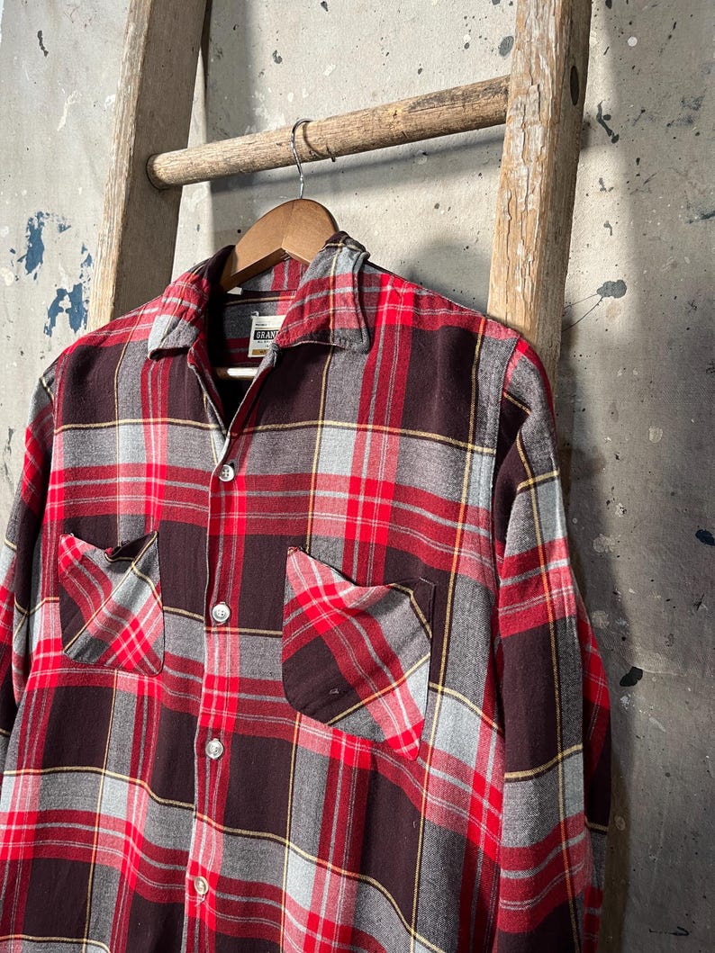 May include: A red, black, and gray plaid flannel shirt with two chest pockets and button closures. The shirt is hanging on a wooden hanger. The shirt has a classic, vintage style.