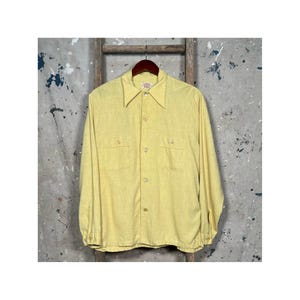 Puede incluir: Una camisa amarilla de manga larga con botones y dos bolsillos en el pecho. La camisa tiene una tela ligeramente texturizada y un cuello clásico.
