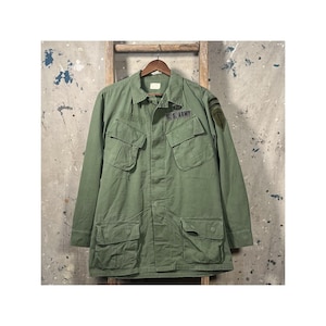 Puede incluir: Chaqueta militar verde oliva con múltiples bolsillos delanteros y cuello. La chaqueta tiene un parche de "U.S. ARMY" y un parche en el hombro. Cuelga de una percha de madera sobre un fondo neutro.