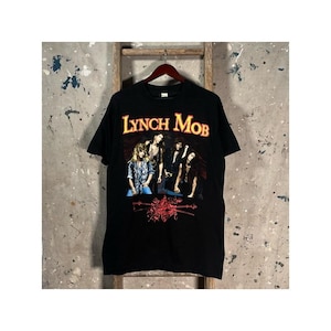 May include: A black short-sleeve t-shirt featuring the text LYNCH MOB in yellow and red lettering above a graphic of a band. Below the band members is a red illustration of a sword and floral elements. The shirt has a crew neckline.