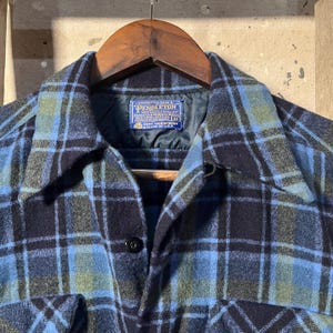 May include: A blue, green, and black plaid wool shirt with a button-down front and a collar. The shirt is hanging on a wooden hanger. A label on the inside collar reads "Pendleton".