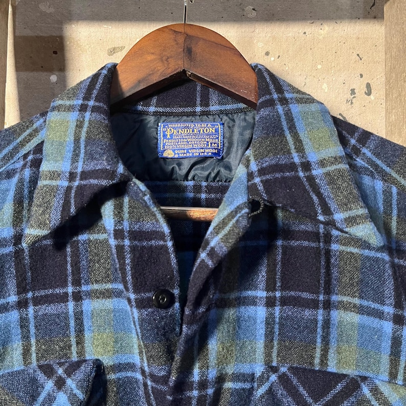 May include: A blue, green, and black plaid wool shirt with a button-down front and a collar. The shirt is hanging on a wooden hanger. A label on the inside collar reads "Pendleton".