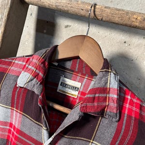 May include: A red, gray, and black plaid flannel shirt with a button-down collar, hanging on a wooden hanger. The shirt has a label that reads "Granella". The shirt is displayed on a wooden ladder.