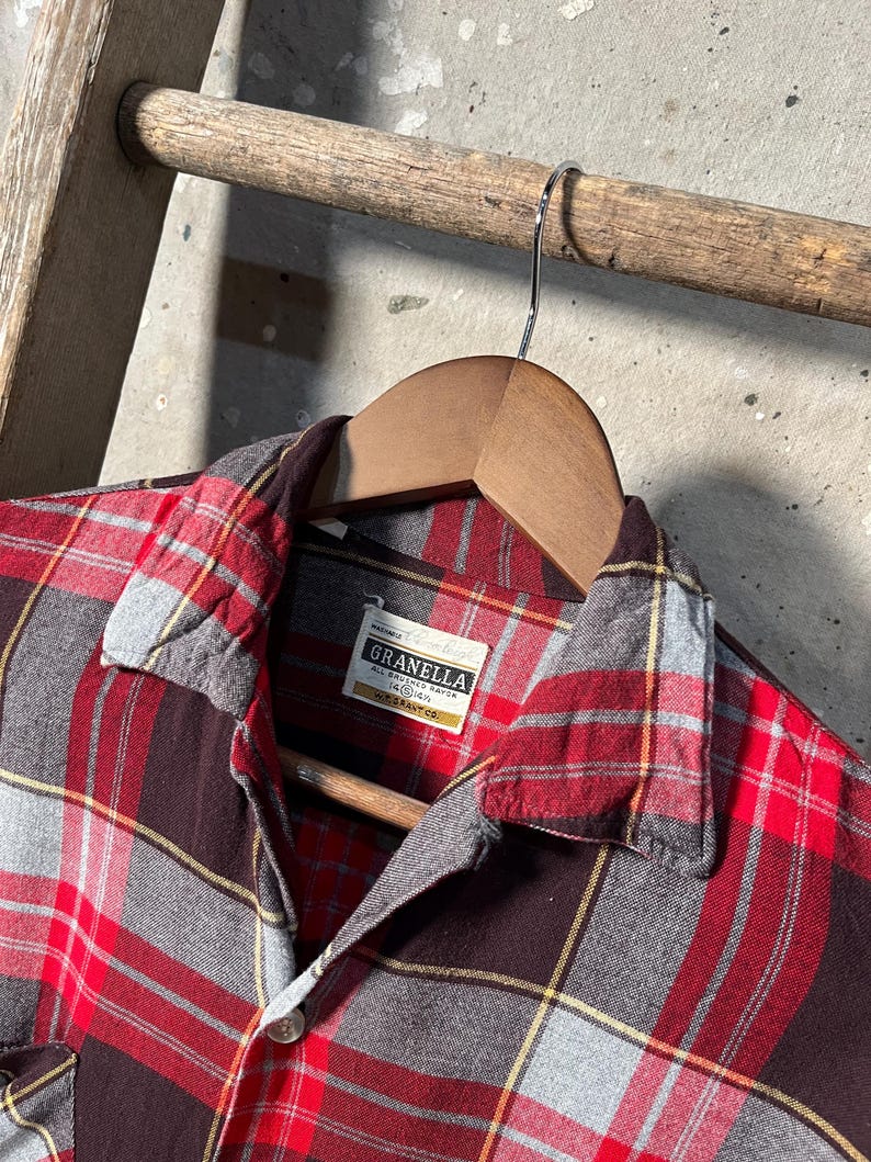 May include: A red, gray, and black plaid flannel shirt with a button-down collar, hanging on a wooden hanger. The shirt has a label that reads "Granella". The shirt is displayed on a wooden ladder.