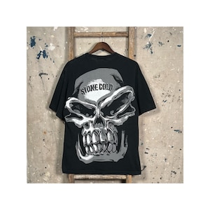 Puede incluir: Camiseta negra con un gran gráfico de calavera en tonos grises y blancos. El texto "STONE COLD" está impreso encima de la calavera. La camiseta está colgada de una percha de madera.