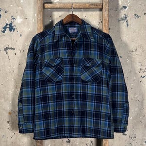 May include: A blue, green, and black plaid button-down shirt with two chest pockets. The shirt is made of a thick, warm fabric, and is hanging on a wooden hanger. The shirt has a classic, vintage style.