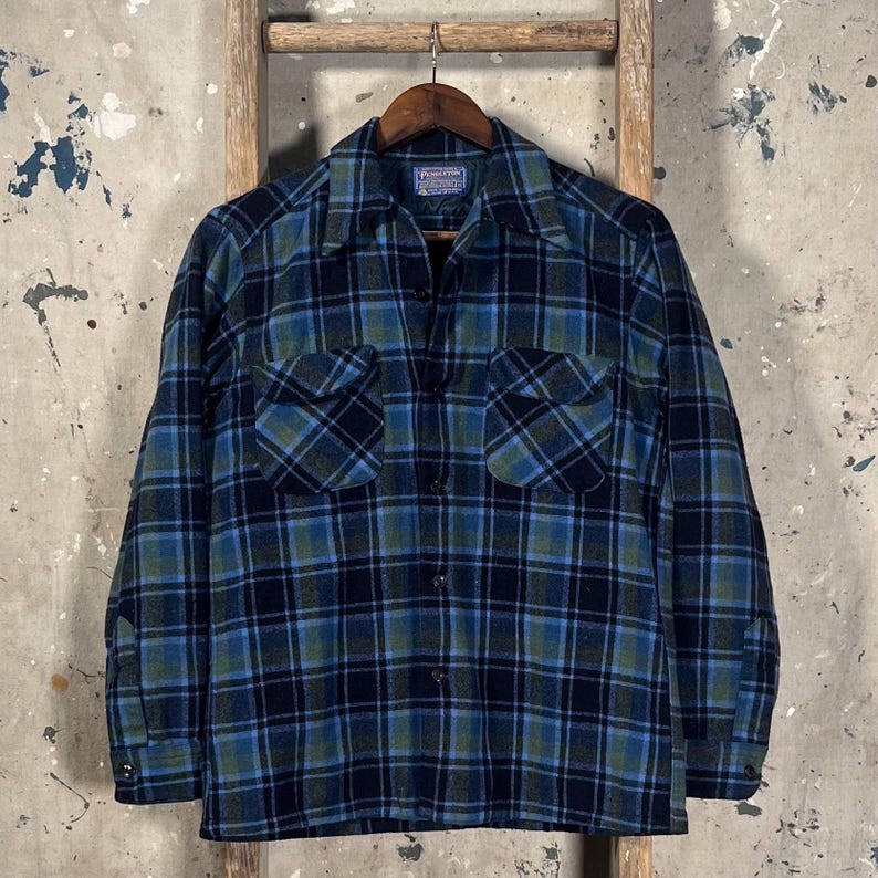 May include: A blue, green, and black plaid button-down shirt with two chest pockets. The shirt is made of a thick, warm fabric, and is hanging on a wooden hanger. The shirt has a classic, vintage style.